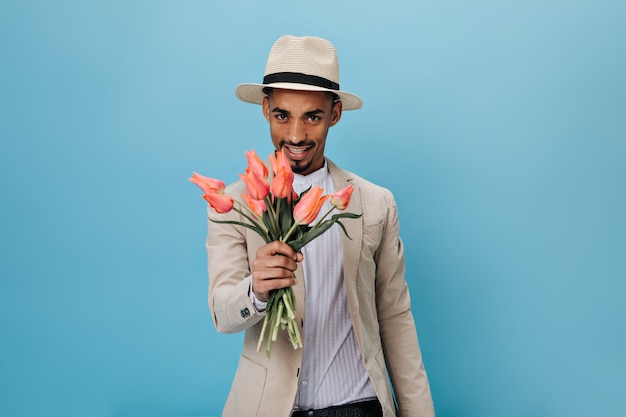 Homme aux yeux bruns au chapeau tenant des tulipes roses sur un mur bleu