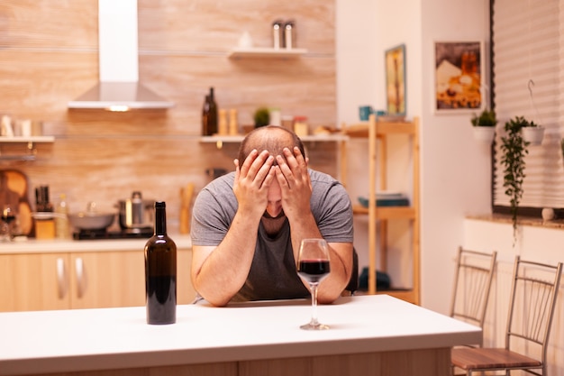 Homme ayant des problèmes d'alcool pendant la crise de la vie avec une bouteille de vin dans la cuisine assis à table. Maladie de la personne malheureuse et anxiété se sentant épuisée par des problèmes d'alcoolisme.