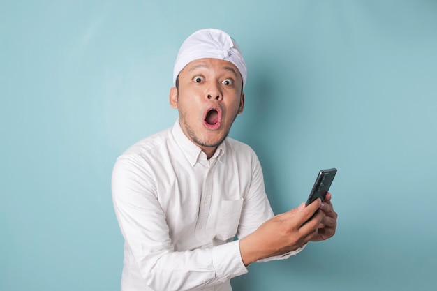 Homme balinais surpris portant un bandeau udeng ou traditionnel et une chemise blanche tenant son smartphone isolé par fond bleu