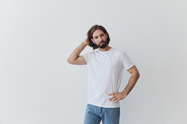 Un homme avec une barbe et des cheveux longs dans un t-shirt blanc et un jean bleu se tient contre un mur blanc appuyé contre lui et écoute de la musique avec des écouteurs blancs sans fil qui regardent pensivement