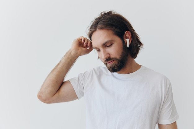 Un homme avec une barbe et des cheveux longs dans un t-shirt blanc et un jean bleu se tient contre un mur blanc appuyé contre lui et écoute de la musique avec des écouteurs blancs sans fil qui regardent pensivement