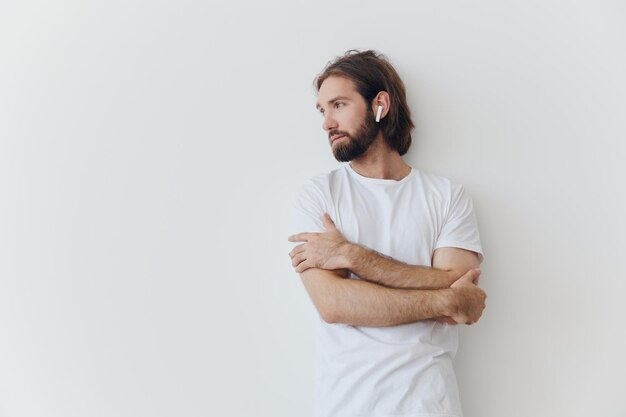 Un homme avec une barbe et des cheveux longs dans un t-shirt blanc et un jean bleu se tient contre un mur blanc appuyé contre lui et écoute de la musique avec des écouteurs blancs sans fil qui regardent pensivement