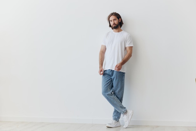 Un homme avec une barbe et des cheveux longs dans un t-shirt blanc et un jean bleu se tient contre un mur blanc penché sur tout son style décontracté