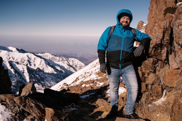 Un homme avec une barbe dans une capuche pose pour la caméra sur fond de montagnes hivernales