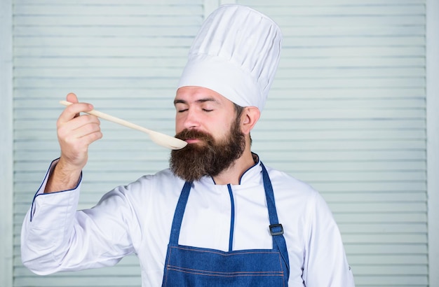 Un homme avec une barbe dans un chapeau de cuisinier et un tablier tient des outils de cuisine La cuisine comme profession professionnelle Le chef barbu hipster tient une cuillère en bois Ustensiles de cuisine et concept de cuisine Essayons de goûter Ajoutez quelques épices