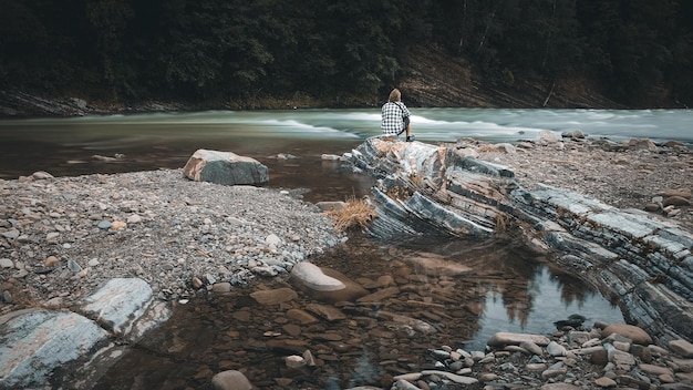 Un homme avec une barbe dans une chemise à carreaux et un short est assis sur la rive d'une rivière de montagne, des couleurs moroses