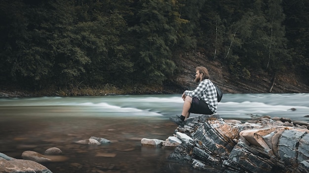 Un homme avec une barbe dans une chemise à carreaux et un short est assis sur la rive d'une rivière de montagne, des couleurs moroses