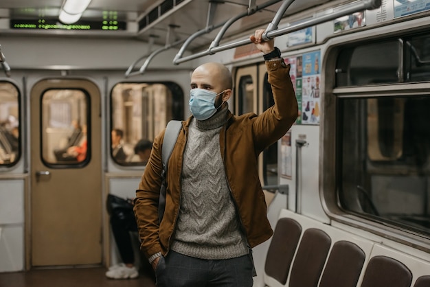 Un homme avec une barbe dans un masque médical pour éviter la propagation du coronavirus tient la main courante dans une voiture de métro. Un gars chauve dans un masque chirurgical contre COVID-19 se tient dans une rame de métro.