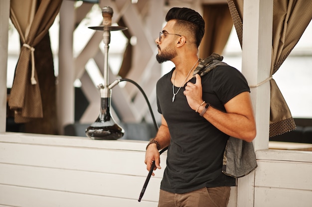 Homme barbe élégant dans des verres et t-shirt noir fumer narguilé en plein air