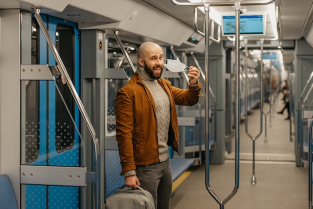 Un homme avec une barbe enlève un masque médical et sourit dans une voiture de métro. Un homme chauve avec un masque chirurgical contre COVID-19 garde une distance sociale dans un train.