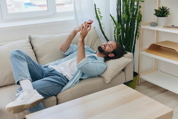 Un homme avec une barbe est allongé sur le canapé pendant la journée à la maison et regarde son téléphone se détendre pendant sa journée de congé un homme jouant à la bourse en ligne sur son téléphone