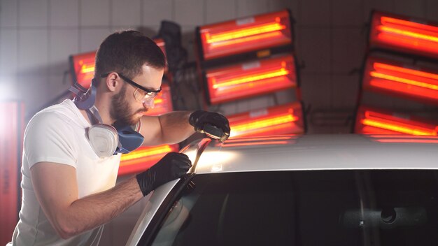Homme avec une barbe et des gants avec une lampe de poche est en train de vérifier la qualité du polissage de la carrosserie de la voiture