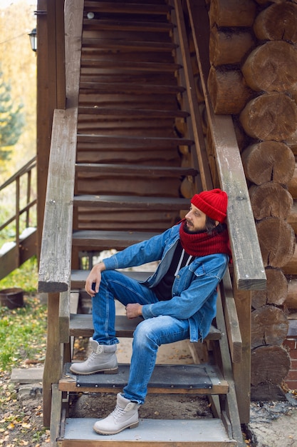 Un homme avec une barbe hipster dans un bonnet rouge et une veste en jean est assis dans les escaliers d'une maison en bois dans les bois