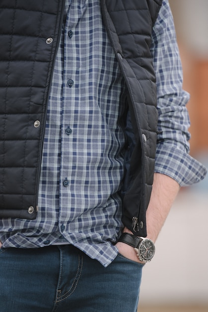 Un homme avec une barbe et des lunettes de soleil dans une chemise à carreaux et un gilet posant dans la rue