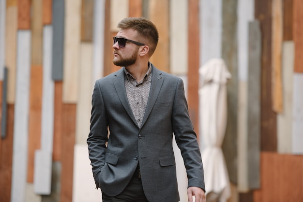 Un homme avec une barbe et des lunettes de soleil dans un costume gris pose dans la rue pour faire de la publicité pour les vêtements pour hommes.