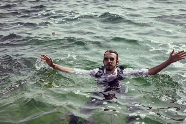 Homme Avec Une Barbe Et Des Lunettes De Soleil En Vêtements Un Gilet Et Une Chemise Blanche Nage En Mer Parmi Les Rochers En été