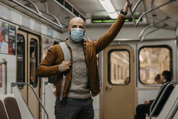 Un homme avec une barbe et un masque médical sur le visage pour éviter la propagation du coronavirus roule dans une voiture de métro. Un homme chauve dans un masque chirurgical contre le COVID-19 se tient dans une rame de métro.