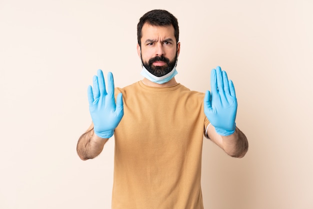 Homme avec barbe protégeant du coronavirus avec un masque et des gants sur un mur isolé faisant un geste d'arrêt et déçu