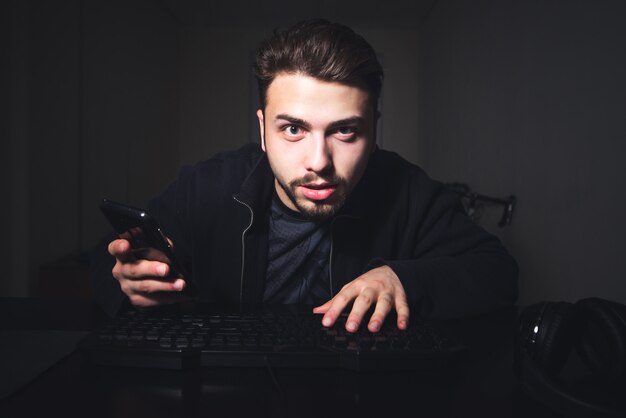 Homme avec une barbe et un smartphone dans ses mains est assis la nuit sur ordinateur