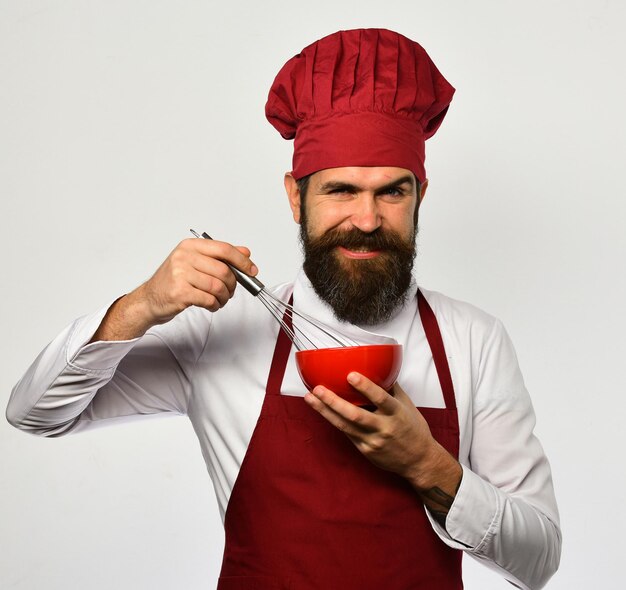 Un homme à la barbe tient des ustensiles de cuisine sur fond blanc Concept de processus de préparation Cuisinier avec un visage souriant en uniforme bordeaux utilise un fouet et un bol Le chef mélange ou fouette les ingrédients dans une assiette rouge