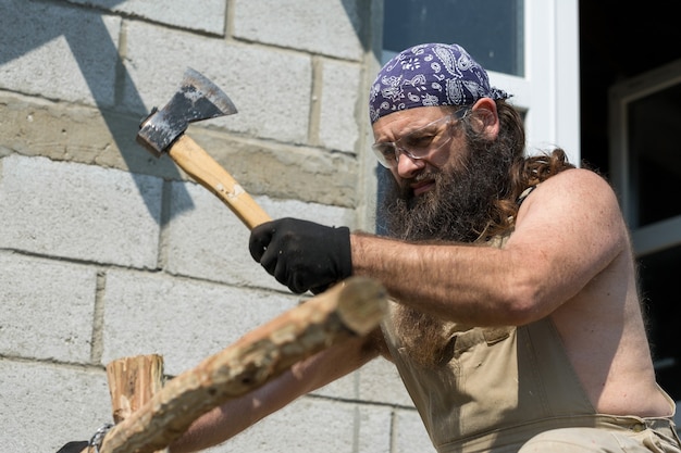 Un homme barbu d'âge moyen dans un bandana coupe des bûches avec une hache Brutal en salopette fait le travail difficile