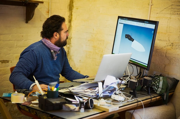 Photo homme barbu assis à un bureau en désordre dans un atelier regardant un écran d'ordinateur