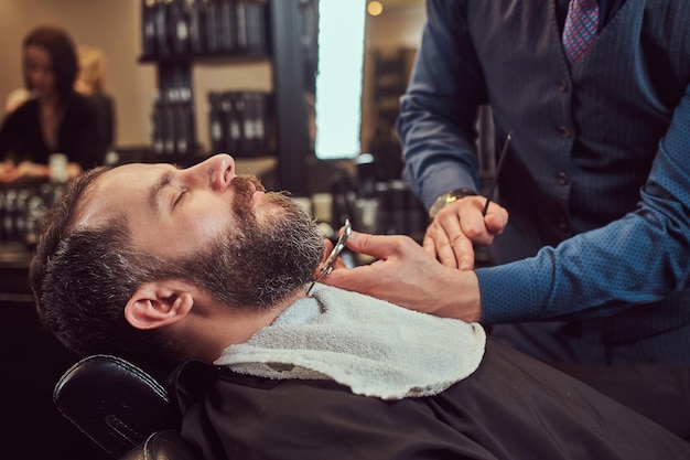 Homme barbu assis dans un fauteuil dans un salon de coiffure pendant que le coiffeur modélise la barbe avec des ciseaux et un peigne au salon de coiffure.