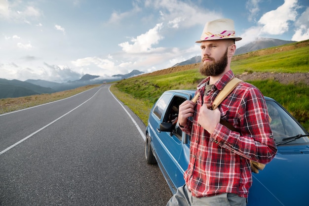 Photo un homme barbu attrayant dans une chemise rouge à carreaux et un chapeau avec un sac à dos se tient à côté d'une voiture rétro