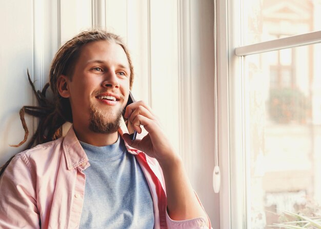 Homme barbu attrayant hipster avec des dreads portant des écouteurs assis près de la fenêtre et à l'aide de smartphone