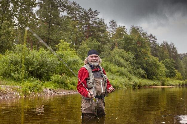 Homme barbu blanc mature attrape du poisson à la mouche dans une rivière calme en forêt, il porte un tissu imperméable, éco-tourisme.