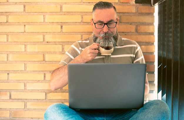 Homme barbu de boire un café tout en regardant l'ordinateur portable assis sur le sol du balcon par un après-midi ensoleillé