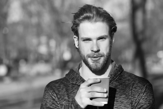 Homme barbu avec une boisson à emporter à l'air frais Macho tient une tasse de café jetable dans un parc ensoleillé Ambiance café ou thé Boisson et nourriture en plein air Thé du matin pour le petit déjeuner