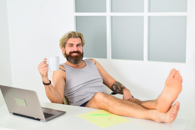 Un homme barbu boit du café et travaille confortablement. auto-éducation en ligne. reste à la maison. verrouiller la quarantaine des coronavirus. entreprise lointaine. homme d'affaires aux pieds nus travaille sur un ordinateur portable dans le bureau à domicile.