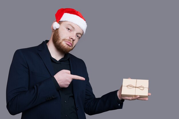 Homme barbu avec bonnet de Noel portant des vêtements de cérémonie pointe vers le présent dans sa main posant sur un mur gris près de l'espace libre