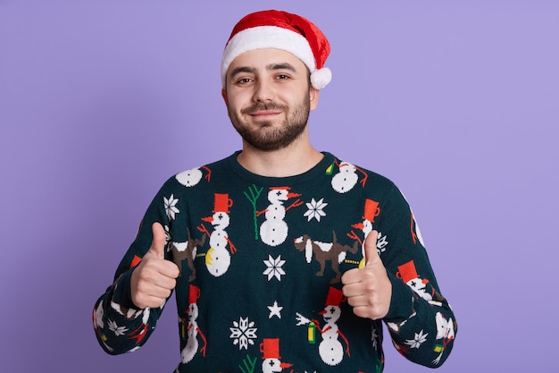 Homme barbu en bonnet de Noel et pull chaud avec des bonhommes de neige montre les pouces vers le haut avec les deux mains