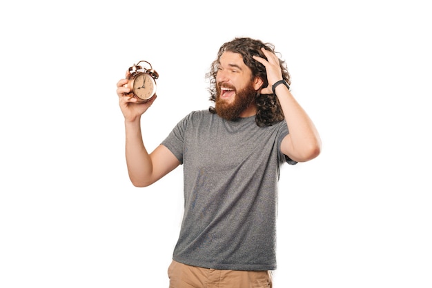 L'homme barbu bouclé surpris regarde un réveil sur fond blanc.