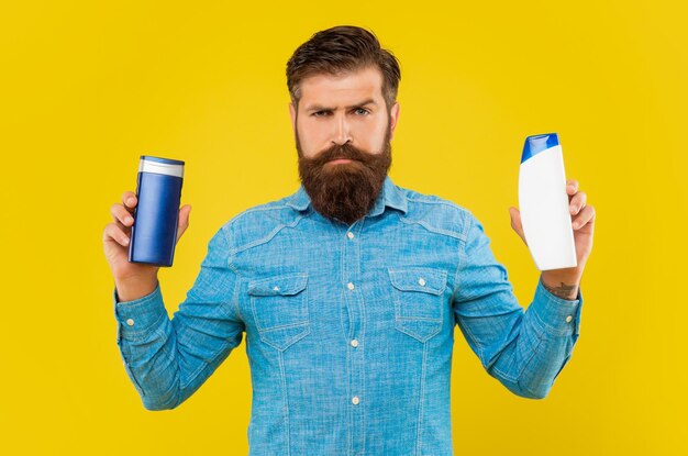 Un homme barbu brutal choisit une bouteille de shampoing sur fond jaune présentant des articles de toilette