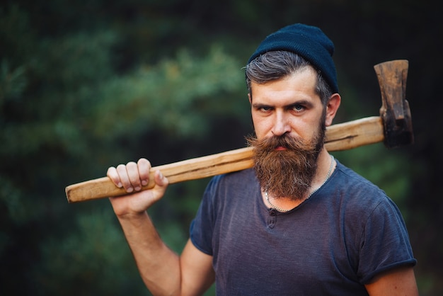 Un homme barbu brutal avec une moustache dans un chapeau chaud et une chemise bleue tient une hache sur son épaule