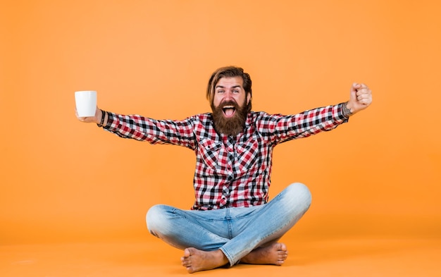 un homme barbu brutal portant une chemise à carreaux ayant une barbe luxuriante et une moustache buvant du café du matin d'une tasse de bonheur
