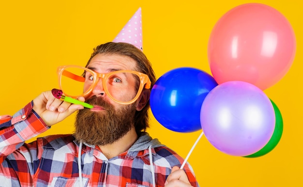 Homme barbu en chapeau de fête et lunettes avec des ballons soufflant la fête d'anniversaire du bruiteur