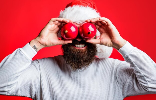 Photo homme barbu avec un chapeau de père noël avec deux jouets pour l'arbre de noël les yeux couverts l'humeur de noël joyeuses vacances