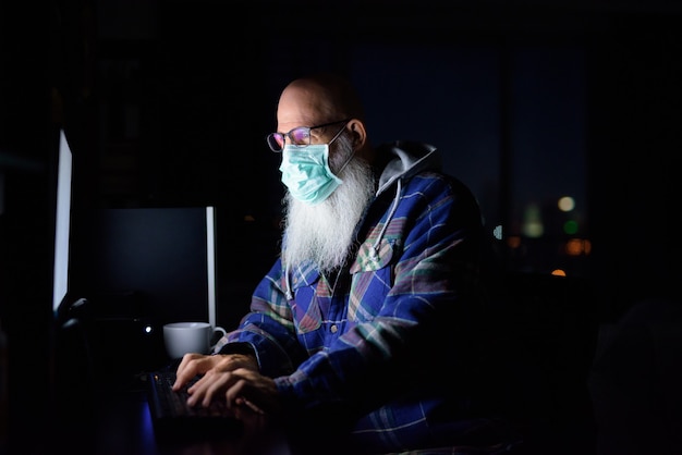 Homme barbu chauve mature avec masque travaillant à domicile dans l'obscurité