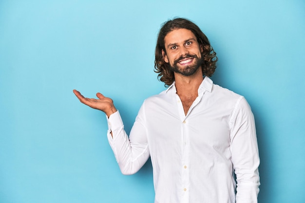 Un homme barbu avec une chemise blanche et un fond bleu montrant un espace de copie sur une paume