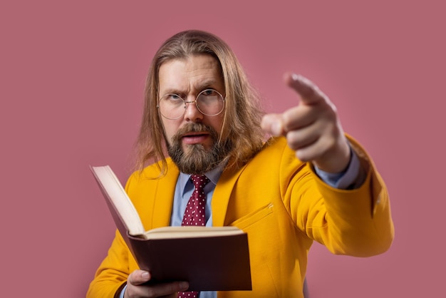 Homme barbu confiant en lunettes et vêtements élégants et lumineux tenant un livre et pointant du doigt sur la caméra Personne mûre avec une expression faciale sérieuse debout en studio avec un fond rose