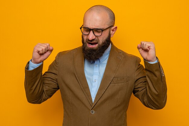 Homme barbu en costume marron portant des lunettes heureux et excité levant les poings se réjouissant de son succès