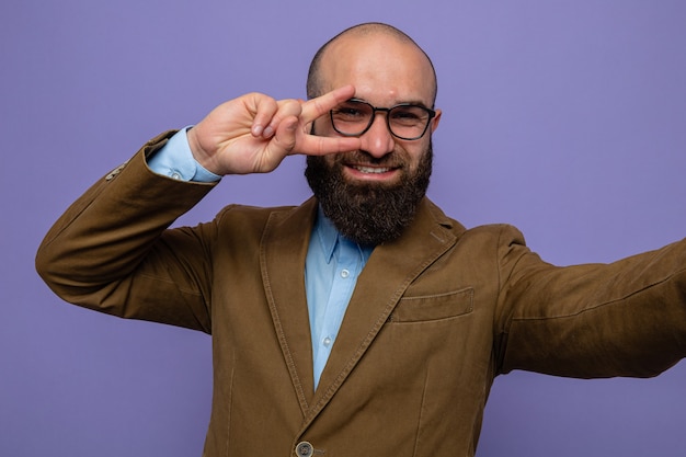 Homme barbu en costume marron portant des lunettes à la prise de selfie souriant montrant gaiement le signe v sur les yeux