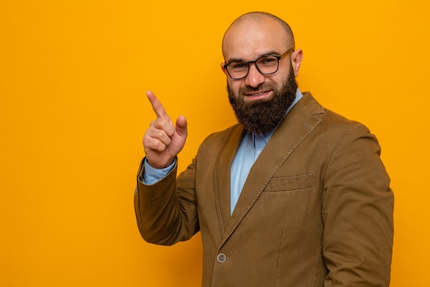 Homme barbu en costume marron portant des lunettes regardant la caméra souriant joyeusement pointant avec l'index sur le côté debout sur fond orange