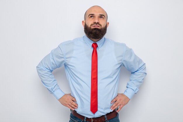 Homme barbu en cravate rouge et chemise bleue jusqu'à perplexe avec les mains à la hanche debout sur fond blanc