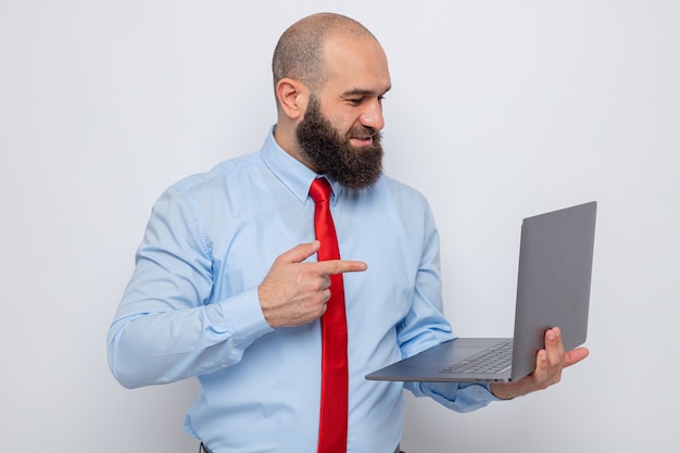 Homme barbu en cravate rouge et chemise bleue tenant un ordinateur portable pointant avec l'index sur l'écran de son ordinateur portable souriant joyeusement debout sur fond blanc