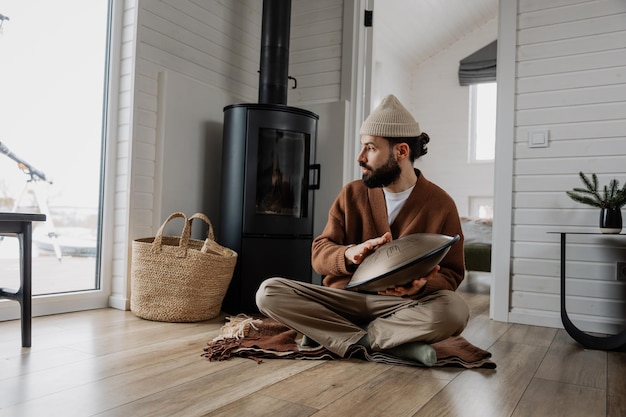 Un homme barbu créatif assis sur le sol à la maison et jouant d'un concept de musique et d'art de tambour accroché